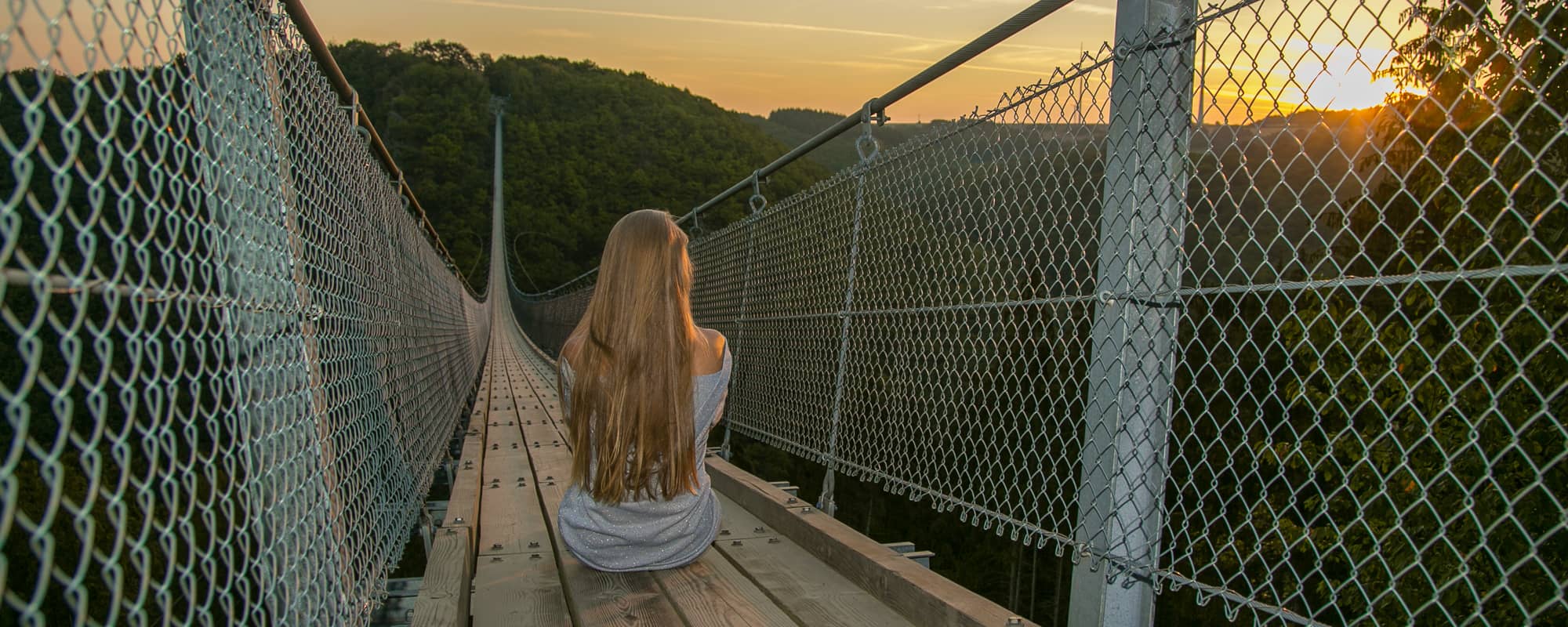 Eine Frau mit langen Haaren sitzt bei Sonnenaufgang auf der Geierlay-Hängeseilbrücke.